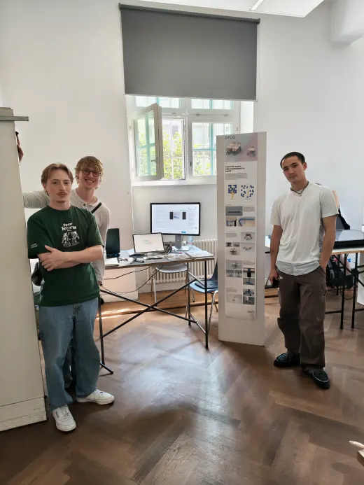 The orca stand at an exhibition: a table with a monitor and a tablet showing the app, a tall poster about the project, and three people standing beside it